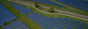 This image shows a large solar farm with rows of solar panels neatly arranged across a grassy field. A pathway runs through the middle of the solar farm, and there are train tracks adjacent to the site. The area is surrounded by greenery and scattered trees.